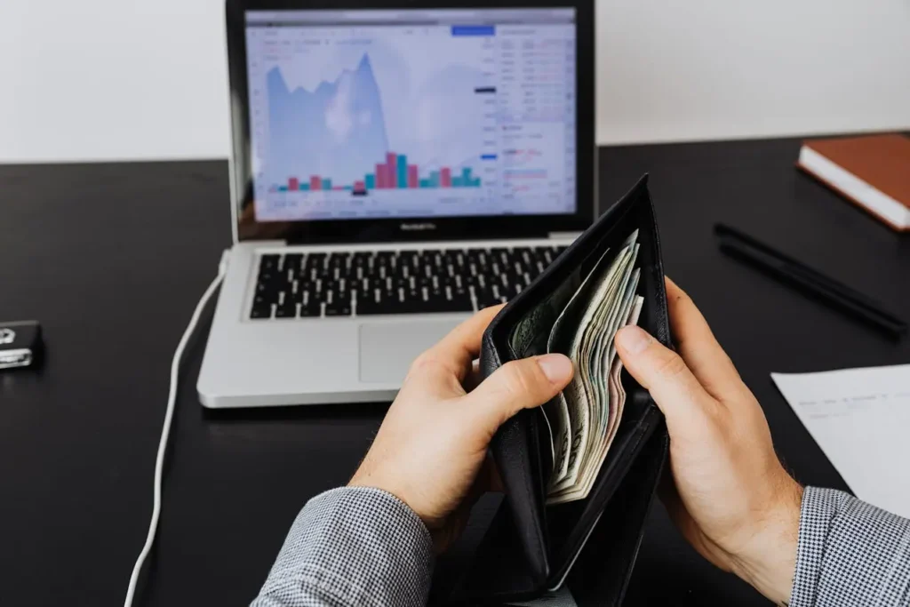 Person holding wallet with cash in front of laptop charts, symbolising budgeting and software development cost planning.
